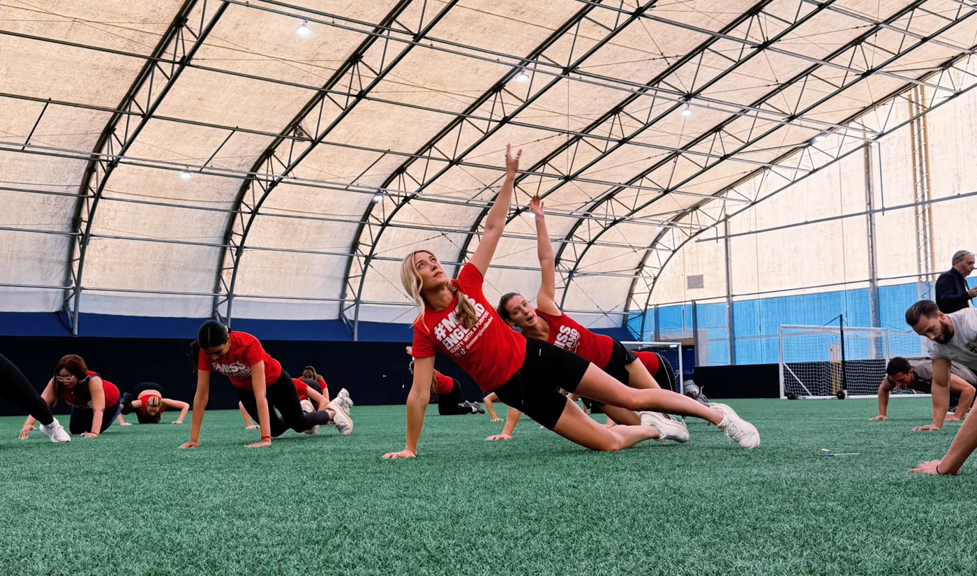 Contestants in red T-shirts stretching sideways with one arm raised on an indoor artificial pitch