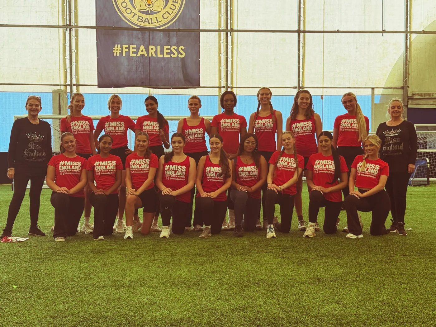 Contestants in red Miss England T-shirts posing in two rows on an indoor pitch beneath a #FEARLESS banner