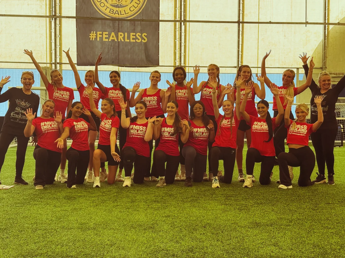 Contestants in red Miss England T-shirts waving at the camera in two rows on an indoor pitch