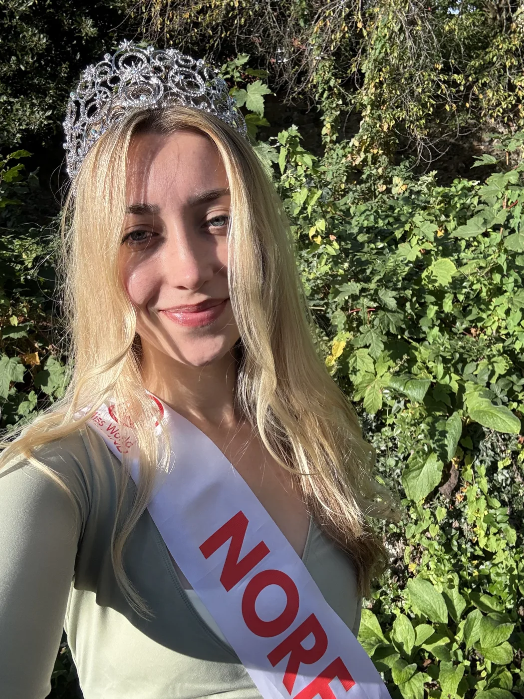 Amy Morris smiling in a crown and a Norfolk sash, with greenery behind her