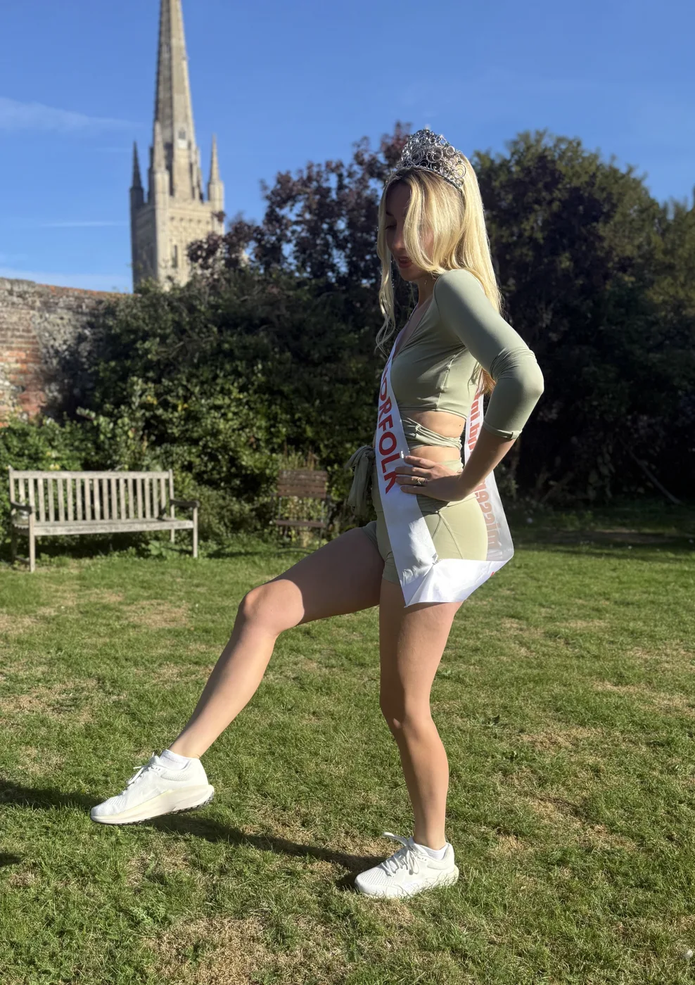 Amy Morris in sportswear, a Norfolk sash and a crown, kicking one trainer forward on a lawn, with a spire and a garden bench behind