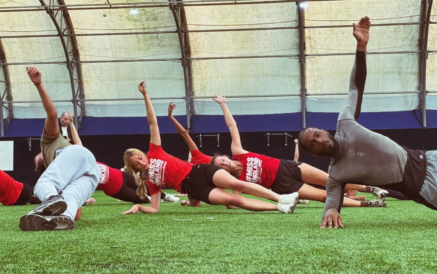 Contestants in red Miss England T-shirts holding side planks on an indoor artificial pitch, with a trainer in grey alongside them