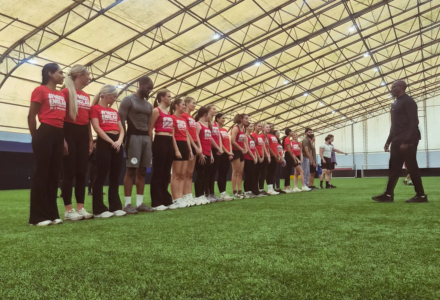 A line of contestants in red Miss England T-shirts on an indoor pitch facing a trainer dressed in black