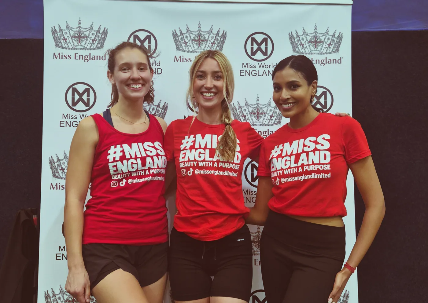 Three contestants in red Miss England tops smiling in front of a Miss England and Miss World England backdrop
