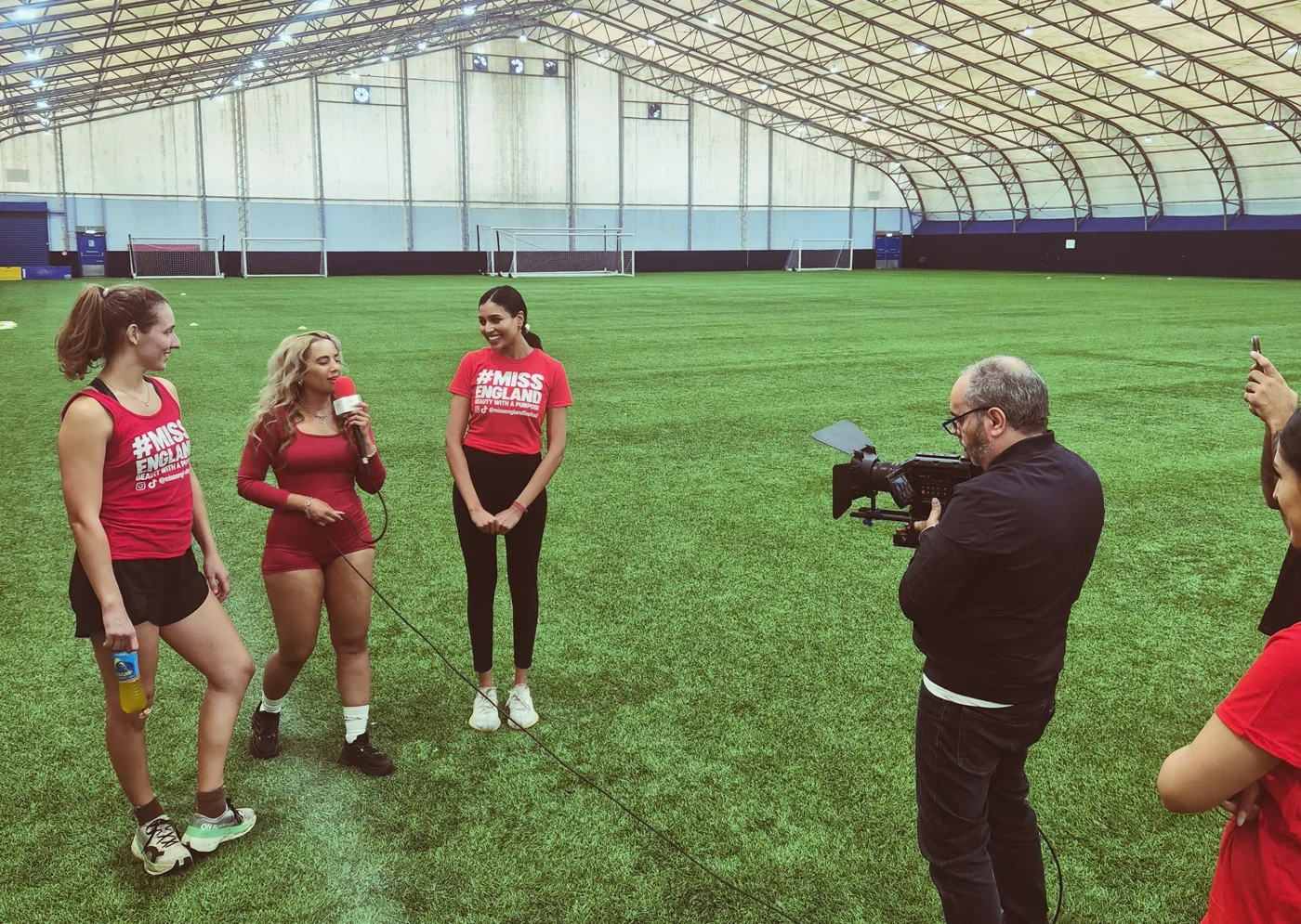 Three contestants being interviewed on an indoor pitch, one holding a microphone, while a man films them on a video camera