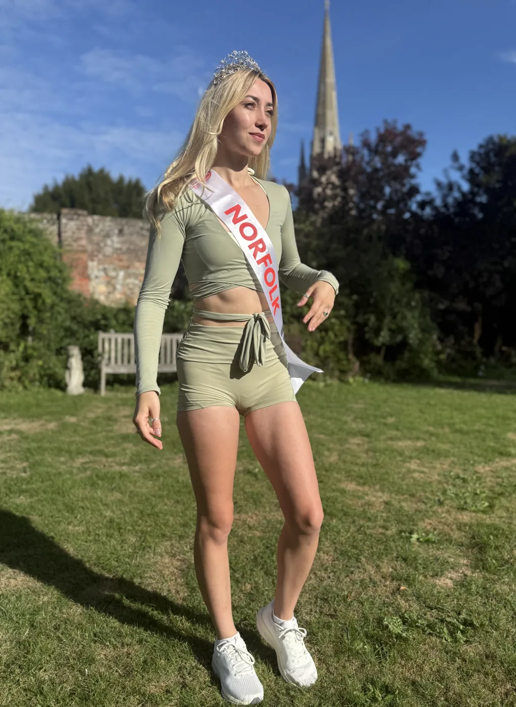 Amy Morris, Miss Norfolk 2026, in her sash, crown and trainers on a lawn with a spire behind