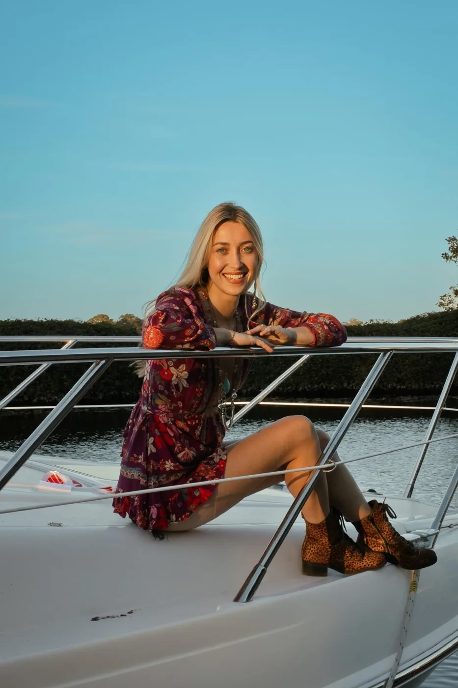 Amy Morris in a red patterned dress, a crown and a Norfolk sash, one arm raised, standing on the deck of a boat under a blue sky