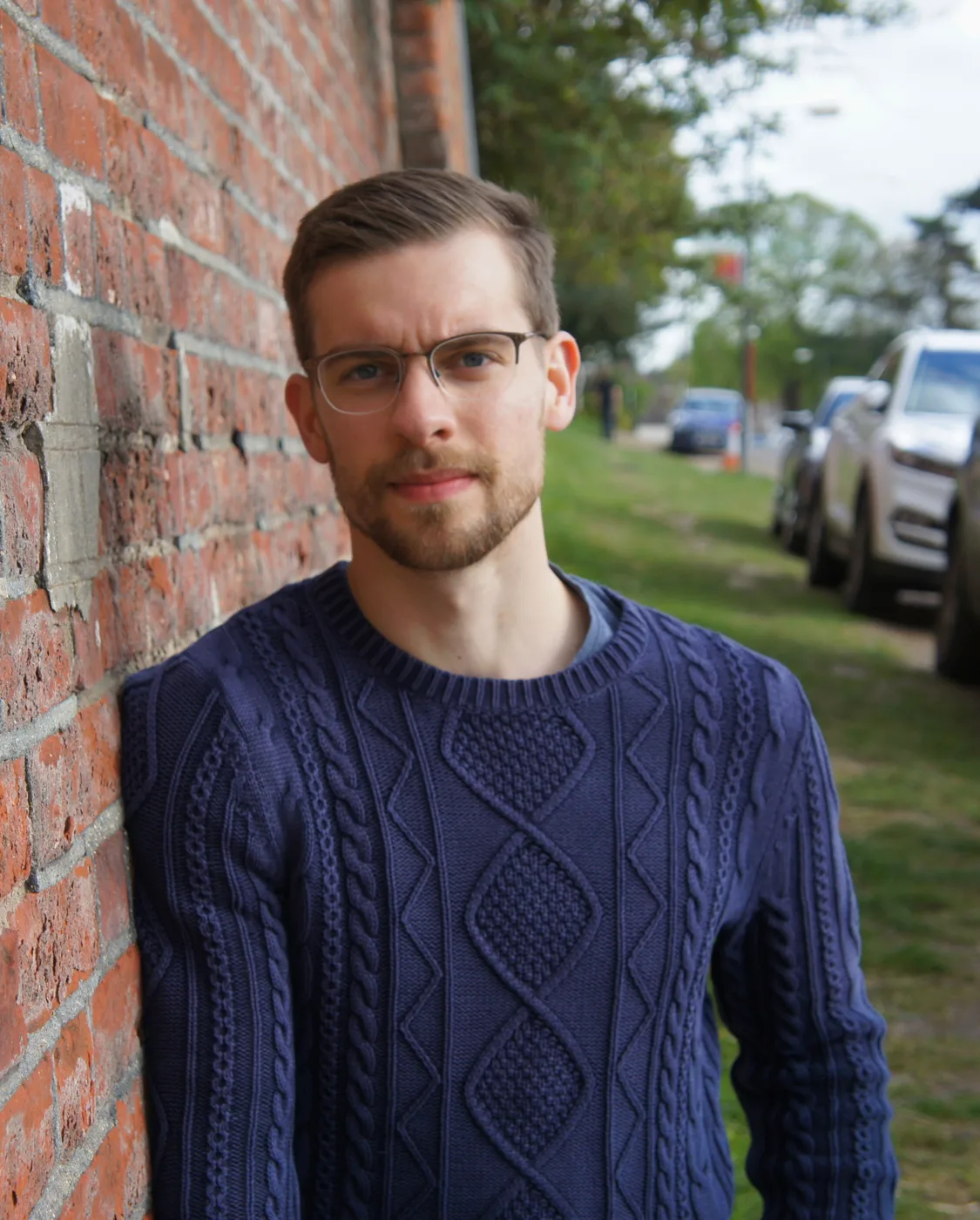 Joe Rejek in glasses and a navy cable-knit jumper, leaning against a red brick wall beside a grass verge and parked cars