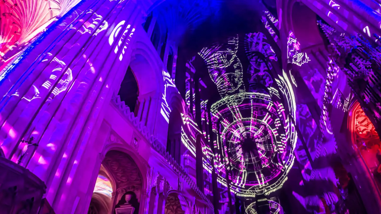 A projected clock mechanism and patterns in purple and blue light on the stone walls and vault of Norwich Cathedral