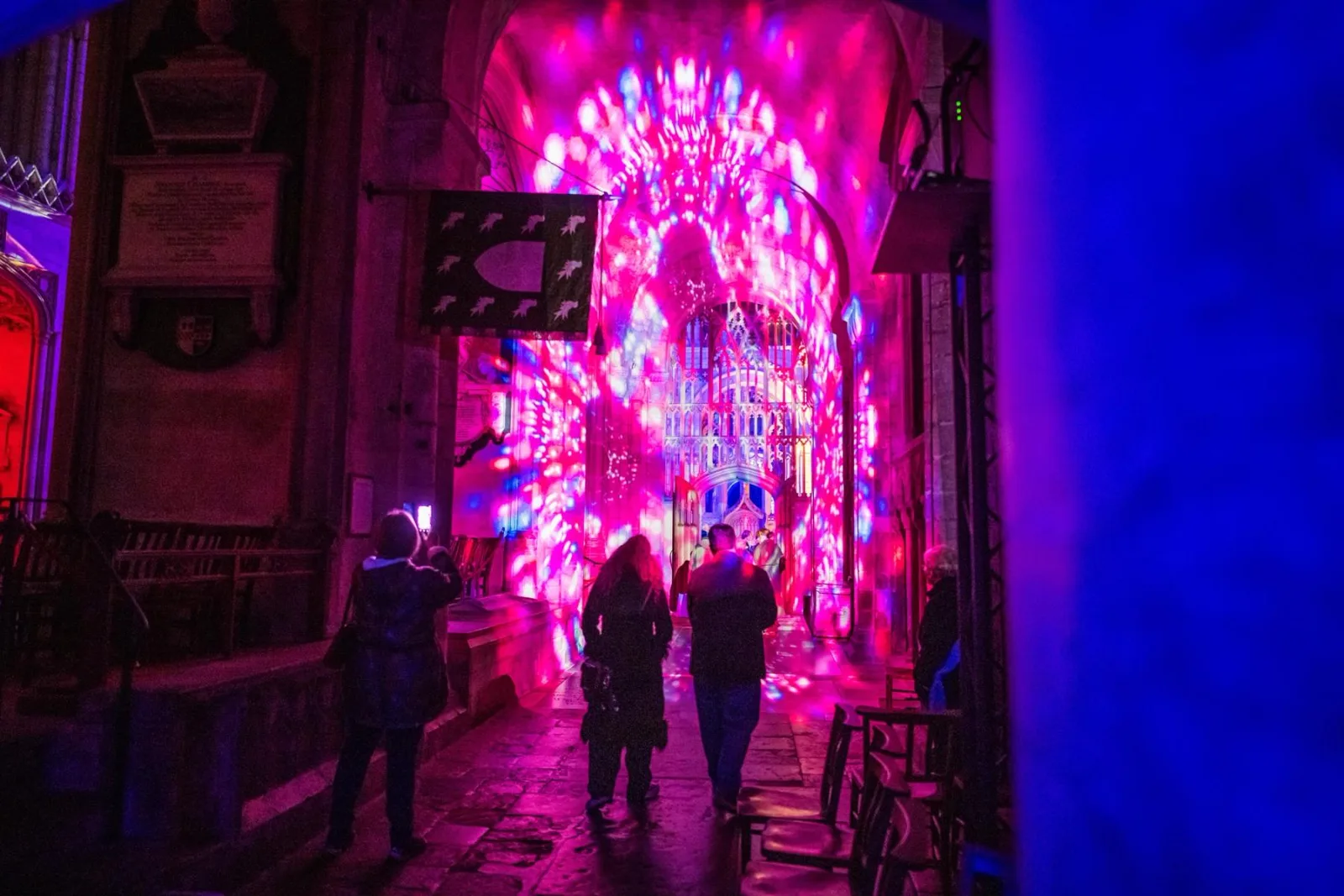 Visitors walk towards a doorway lit in pink and red projected patterns inside Norwich Cathedral