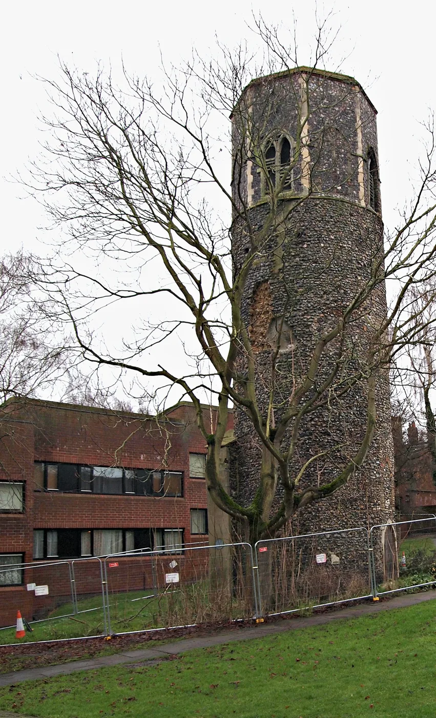 The round flint tower of St Benedict's church in Norwich, with an octagonal top stage, beside a block of flats and a line of temporary fencing